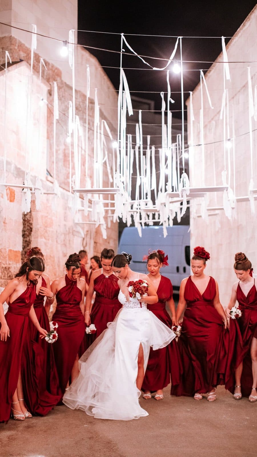 Sposa e damigelle in rosso sotto un'installazione di nastri bianchi in un vicolo di pietra